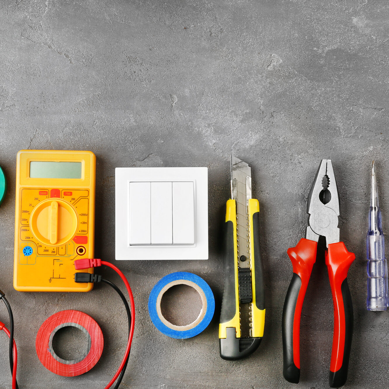 An assortment of electrical tools on a gray surface, including a multimeter, a white light switch, a yellow utility knife, a pair of pliers with red and black handles, a screwdriver with a transparent handle, and rolls of red, blue, and green electrical tape.