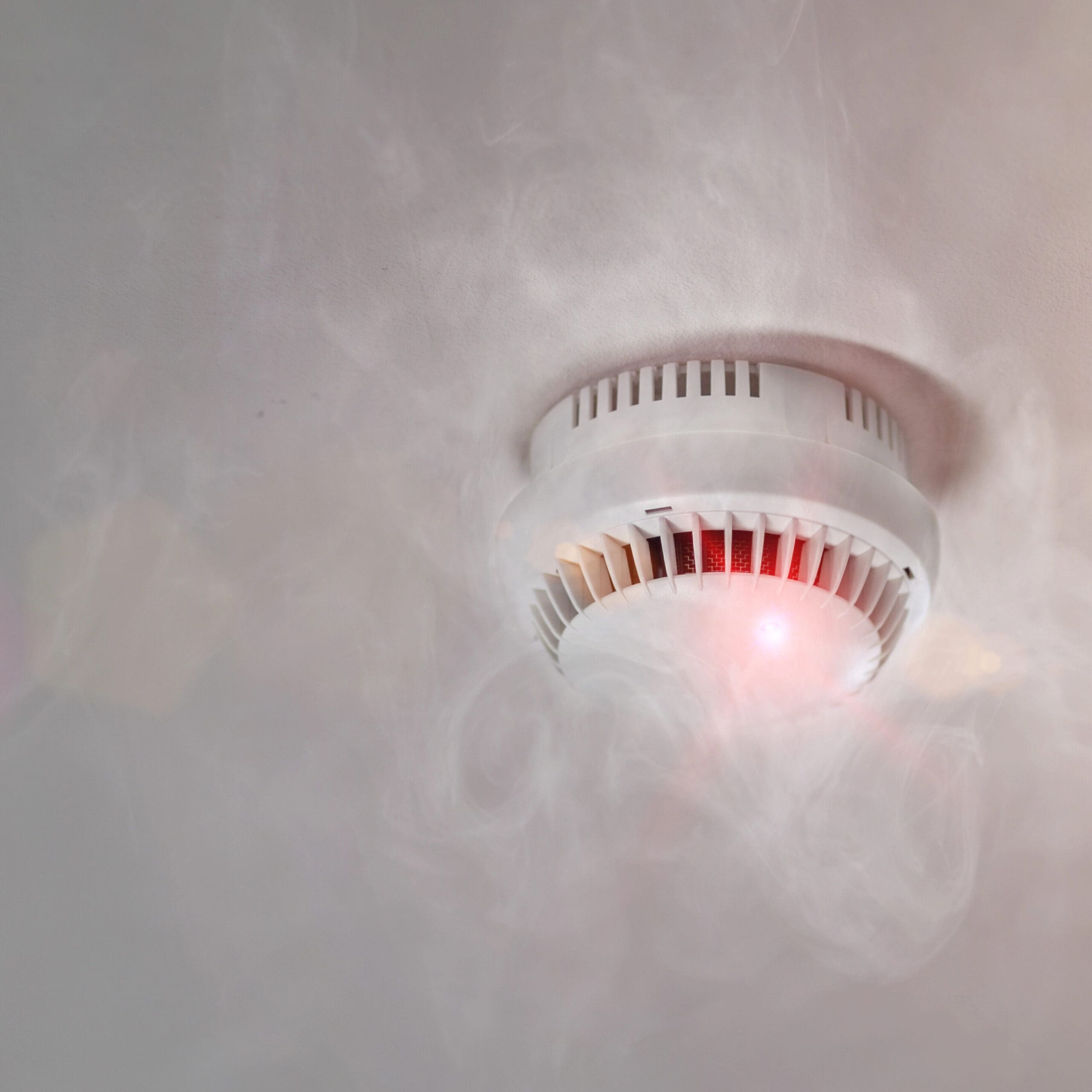 A ceiling-mounted smoke detector with a red light is emitting smoke, indicating that it is activated. The surrounding area is hazy with visible smoke, emphasizing the potential presence of a fire hazard.