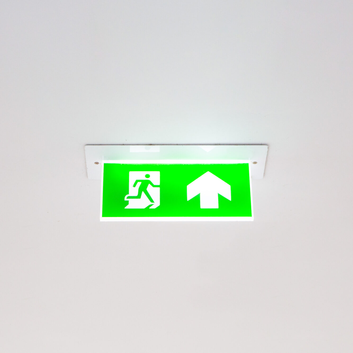 Green exit sign mounted on a ceiling, featuring a white pictogram of a person running through a door on the left and an upward-pointing arrow on the right, indicating the direction of an emergency exit route.