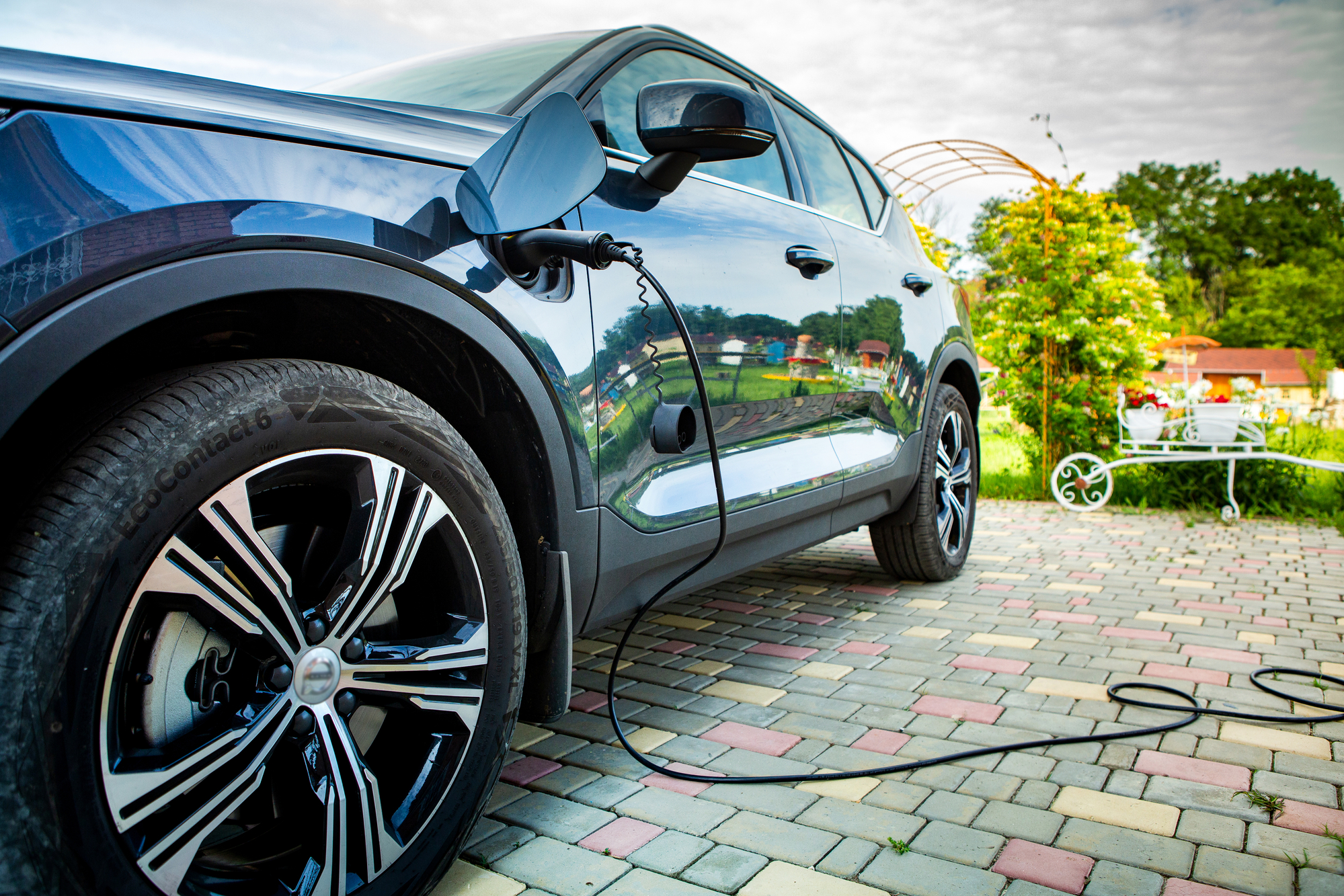 A black electric vehicle is parked on a cobblestone driveway, plugged into a charging station. The background features a garden with greenery, a white bench, and a glimpse of a building. The sky is partly cloudy.