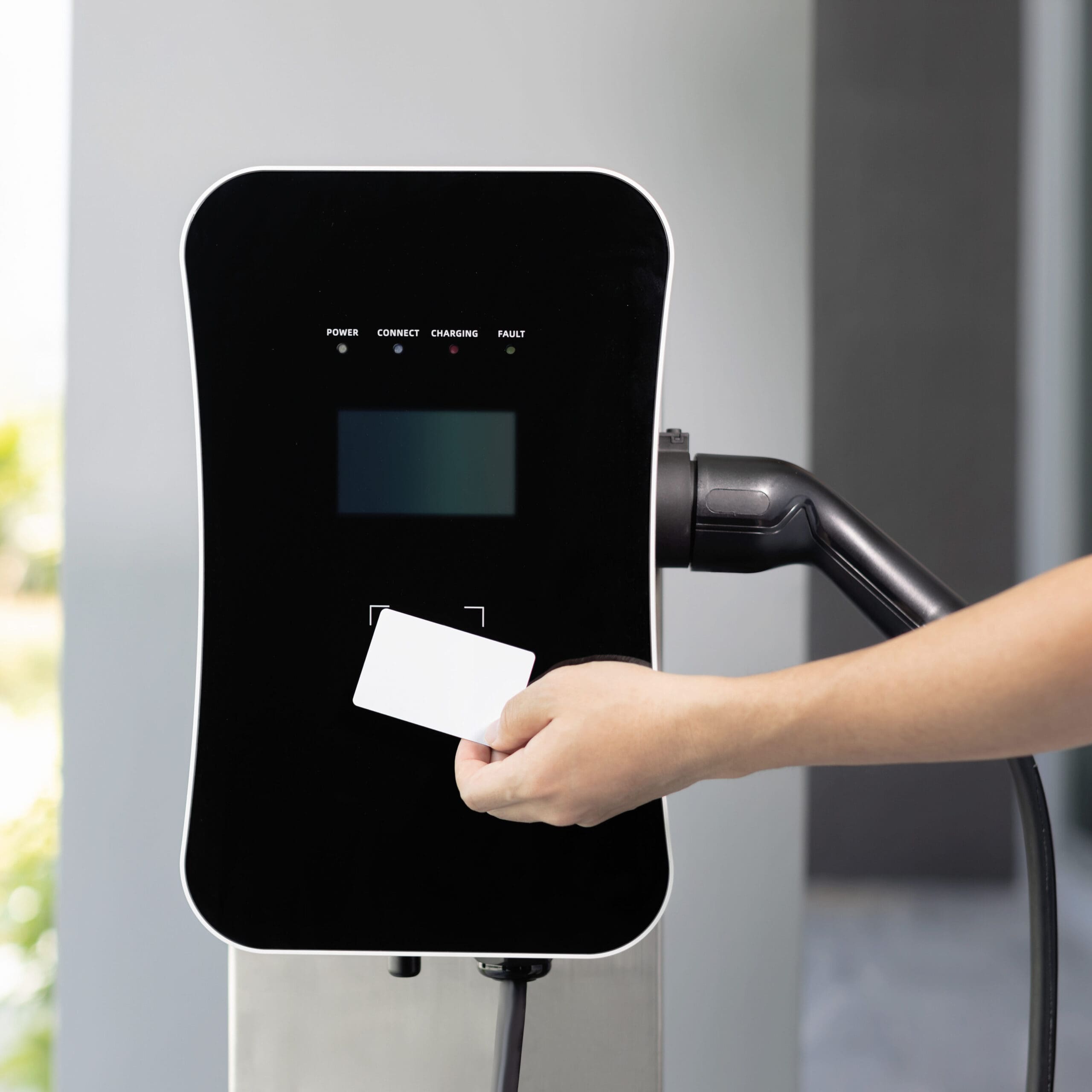 A person holds a white card up to an electric vehicle charging station, initiating a charging session. The station's screen displays indicators for power, connect, charging, and fault statuses. A charging cable is plugged into the station on the right.