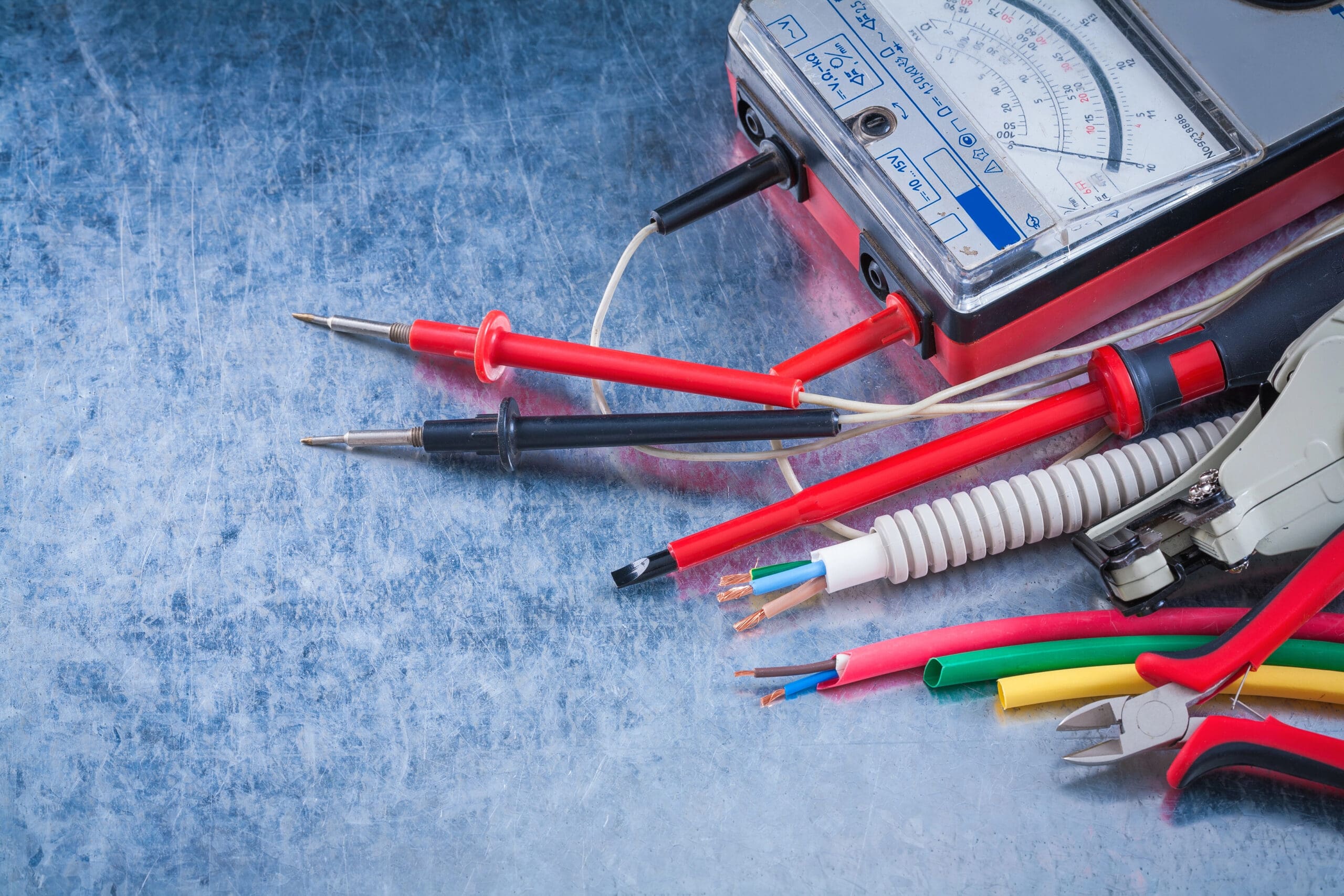 A cluttered workspace featuring a multimeter with red and black probes, various electrical wires of different colors and thicknesses, and a pair of wire cutters. The background has a textured, metallic surface.