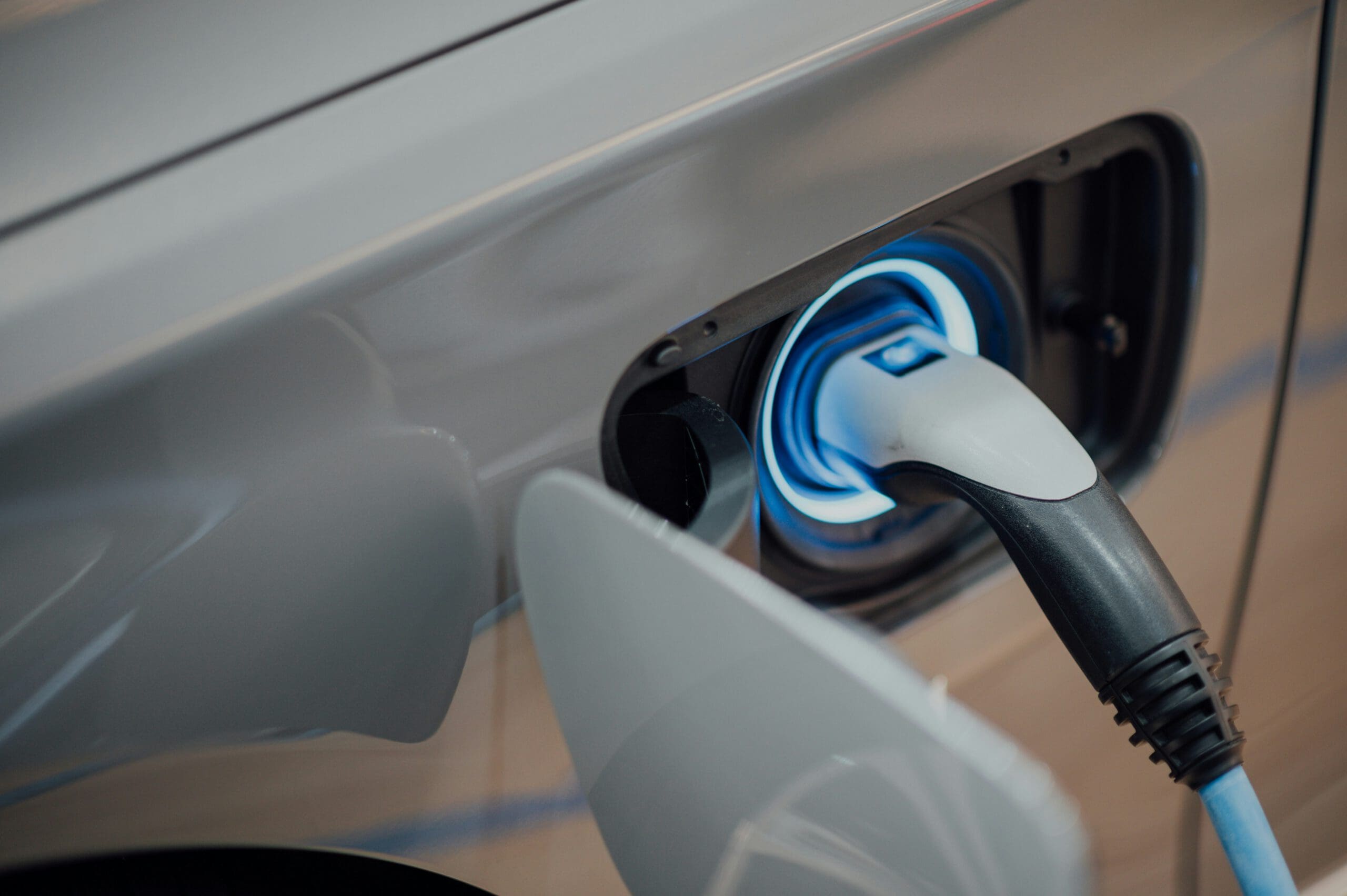 Close-up of an electric car being charged. The charging plug is inserted into the vehicle’s port, which is illuminated with a soft blue light. The car's grey exterior is partially visible.