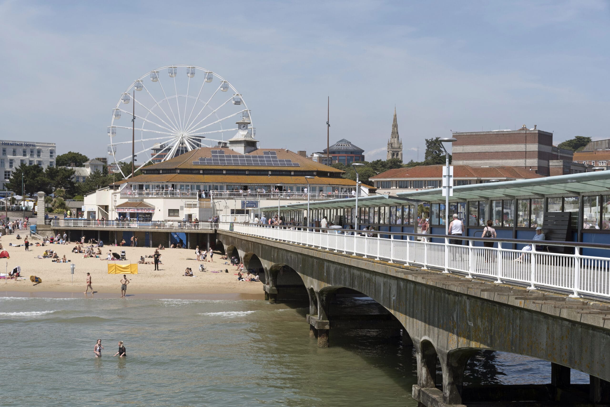 A coastal scene features a bustling beach with people enjoying the sun and water. An amusement park with a Ferris wheel is visible in the background, along with various buildings. In the foreground, a pier extends over the water with people walking on it.