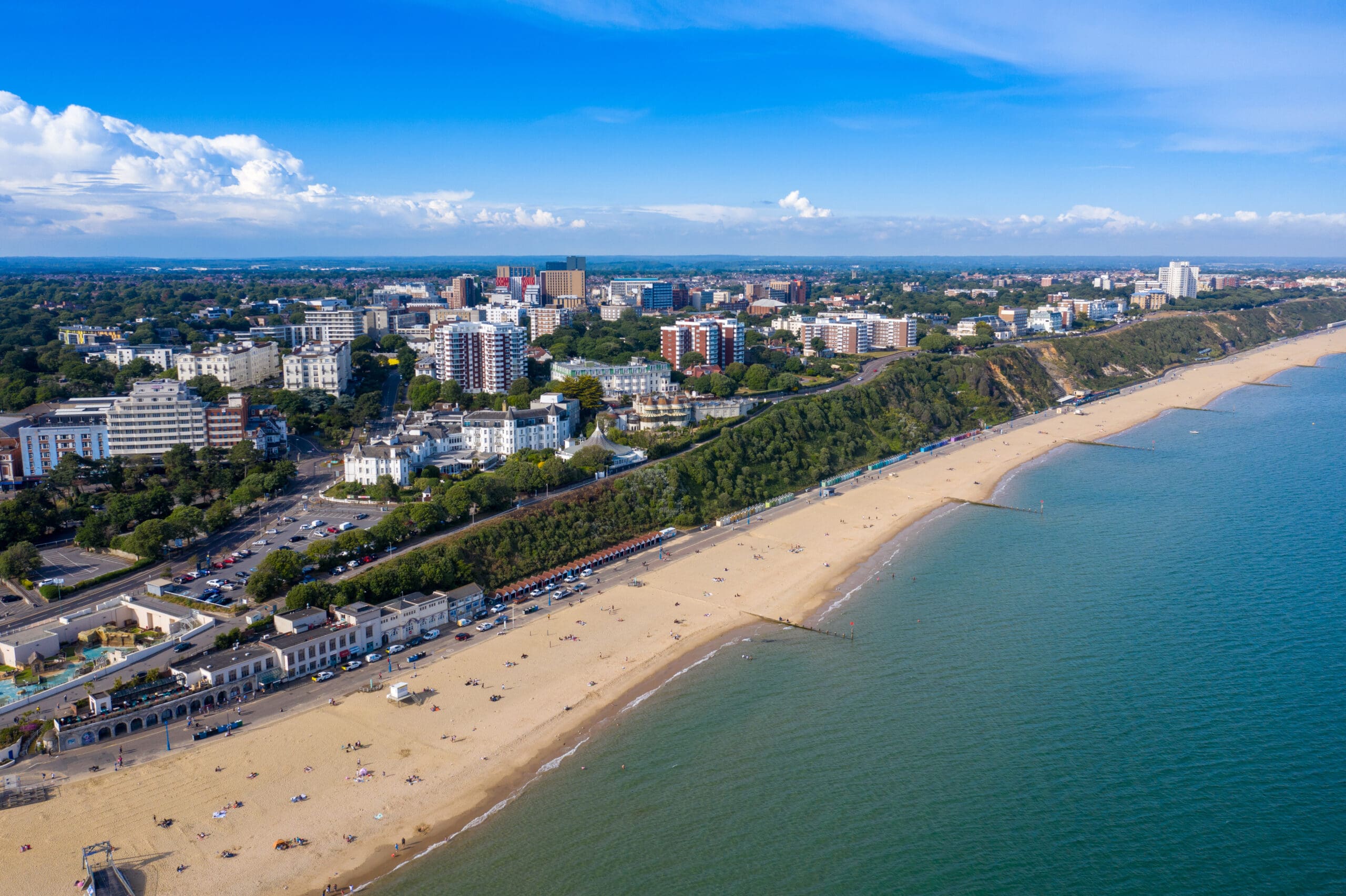 Aerial view of a coastal city with a long sandy beach stretching along the shore. The beach is peppered with people and various structures. The cityscape in the background features numerous buildings, greenery, and the sea is calm with clear blue skies above.