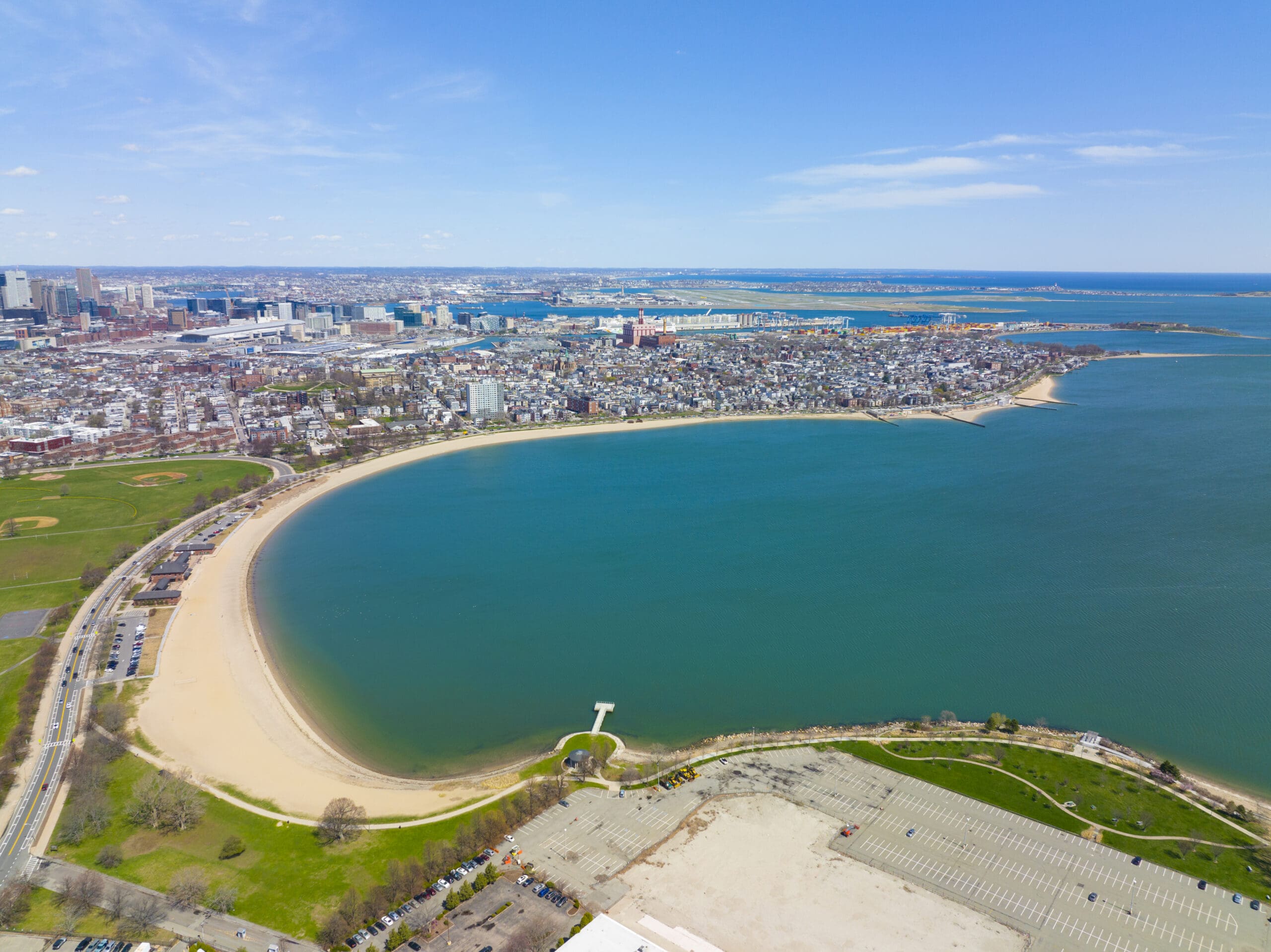 Aerial view of a coastal city with a prominent curved sandy beach along the shoreline. There are residential and commercial buildings in the background, bordered by the ocean. A parking lot and greenery are visible in the foreground. The sky is clear and blue.