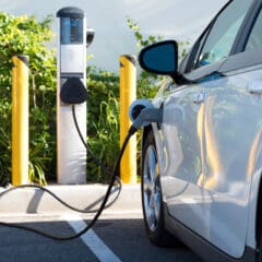 A white electric car is plugged into a charging station in an outdoor parking lot. The charging station is between four yellow bollards, with green foliage and a white background behind it. The car is connected with a cable to the station.