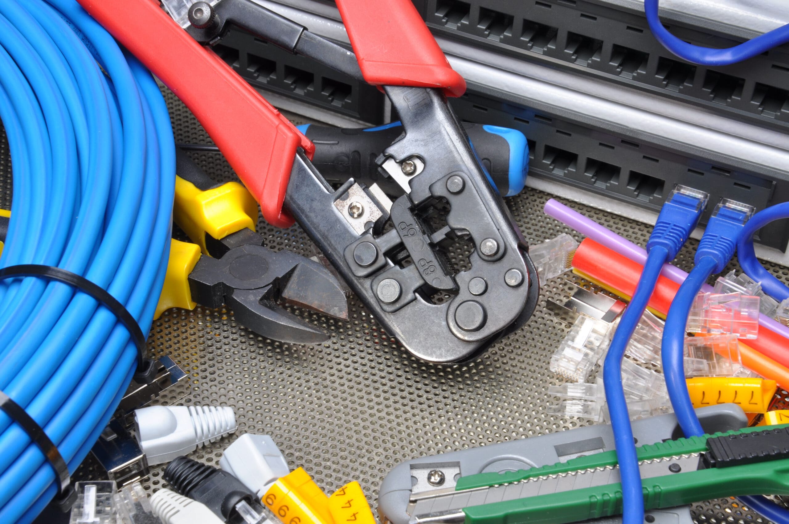 A collection of network tools and accessories including a crimping tool with red handles, wire cutters, blue Ethernet cables, cable ties, and network jacks are scattered on a perforated metal surface.