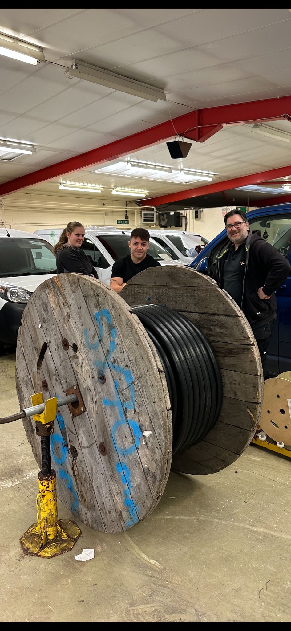 Three individuals stand in a workshop next to a large wooden spool of cable. The person on the left leans on the spool, while the individuals in the middle and to the right pose with smiles. Vans and various equipment are visible in the background.