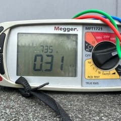 A digital Megger MFT1721 multimeter is displayed, with red, blue, and green test leads attached. The screen shows a reading of 735 A and 0.31 Ω. The device is placed on a concrete surface, next to a blue edge, with a gray background behind it.