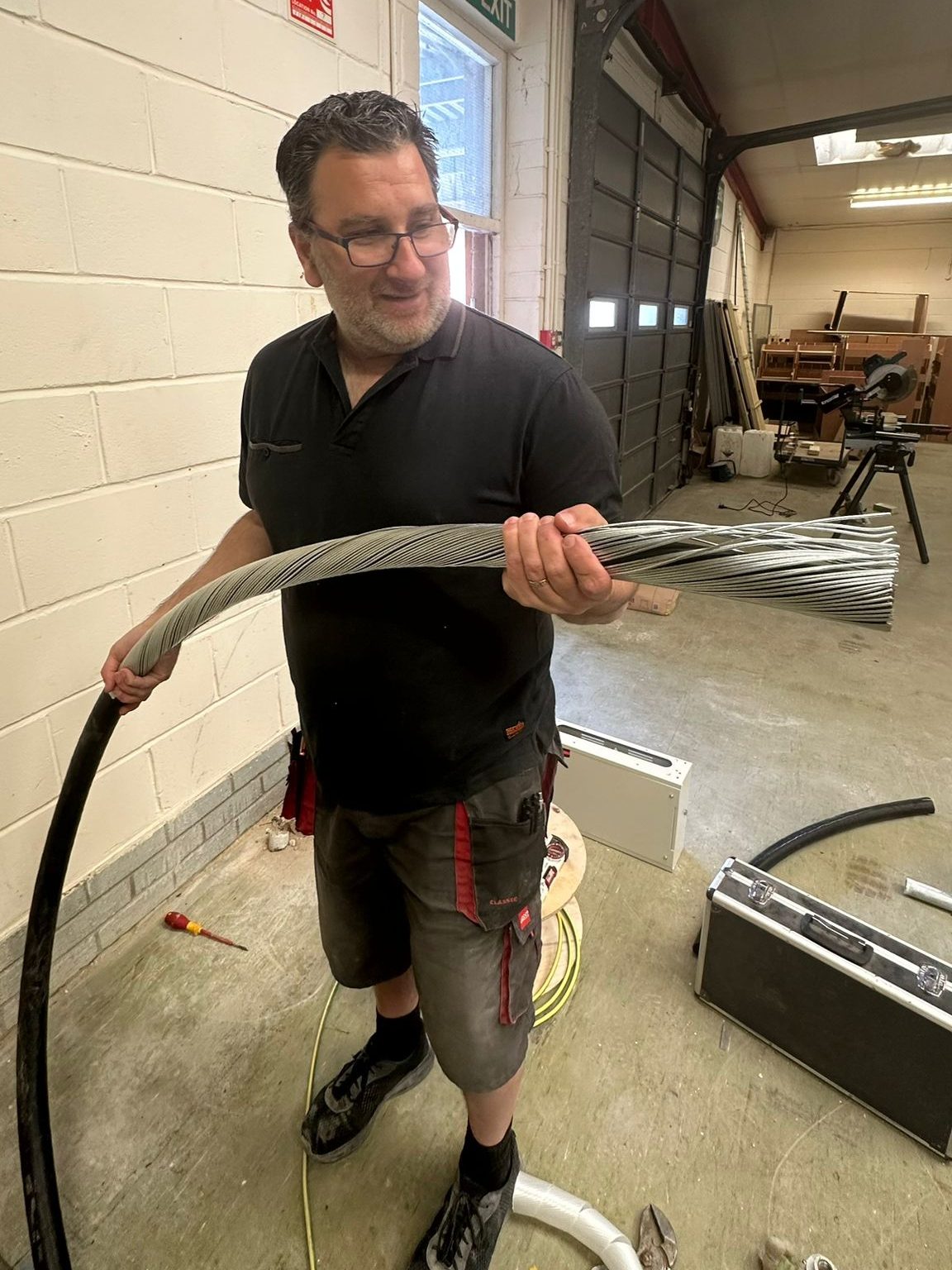 A man in a workshop is holding a thick bundle of wire. He is dressed in a black polo shirt and grey shorts. The workshop background includes various tools and equipment. The man is looking at the wire and smiling slightly.