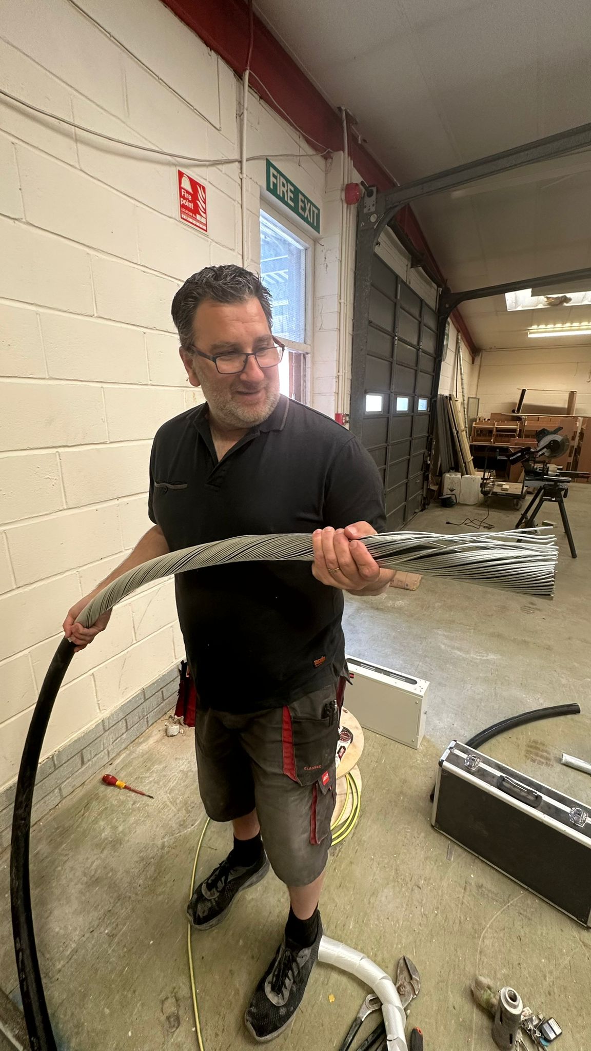 A person wearing glasses and a black shirt, holding a large bundle of thick cables in a workshop. The workshop has various tools and equipment around, with a door and a "Fire Exit" sign visible in the background.