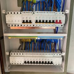 A neatly organized electrical panel with circuit breakers. The panel features two main rows filled with switches, surrounded by blue, yellow, green, and brown wires, each connected to designated terminals. The background shows a plain wall and copper pipes.