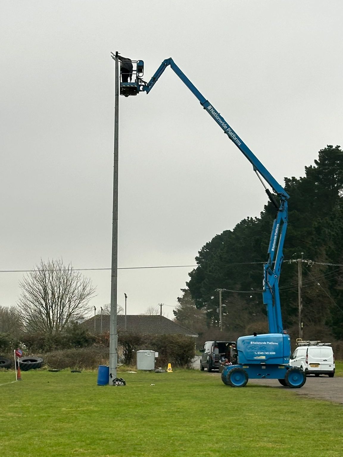 A person is elevated on a blue cherry picker, working on a tall light pole. The cherry picker is positioned on a grassy field with trees, several vehicles, and a building in the background. The sky is overcast.