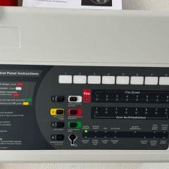 A fire alarm control panel with labeled buttons and indicator lights, showing instructions and various fire zones. The panel includes options for activating and resetting alarms, as well as indicators for power, fire, and fault statuses. Multiple fire zones are listed.