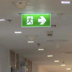 A ceiling-mounted emergency exit sign with a green background and white pictogram showing a person running toward a right-facing arrow. The sign is well-lit, indicating the direction to the nearest exit. Ceiling lights and part of a corridor are visible.