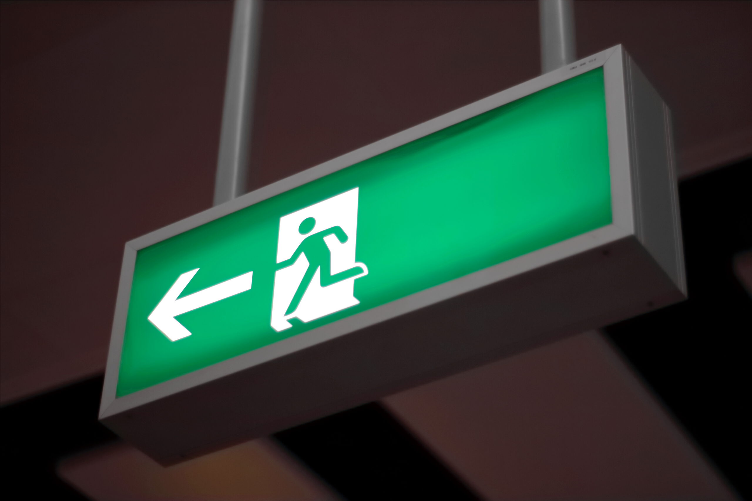 A green emergency exit sign with a white running figure and an arrow pointing left, indicating the direction to the nearest exit. The sign is suspended from the ceiling.