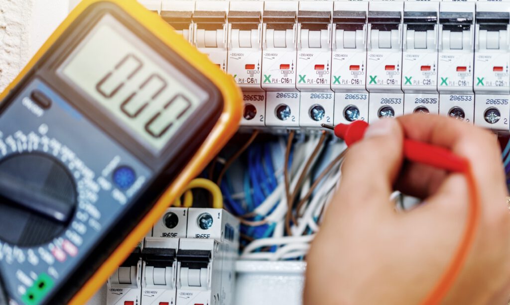 A hand uses a red probe to test wires in an electrical panel, while a digital multimeter displays a reading of 0.00.