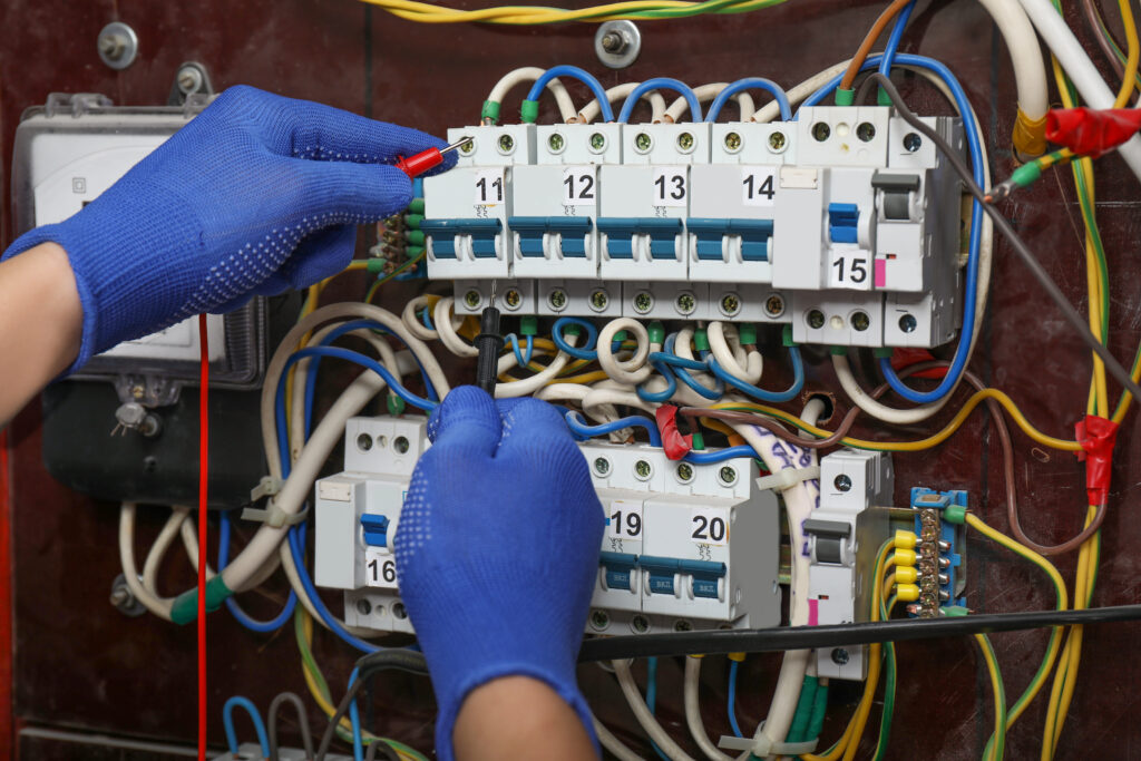A person wearing blue gloves uses probes to test electrical connections in an open circuit breaker panel with multiple labeled switches and many colorful wires.
