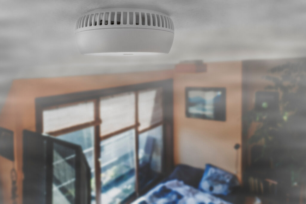 A white smoke detector on the ceiling of a room filled with smoke, with furniture, windows, and an open door visible below.