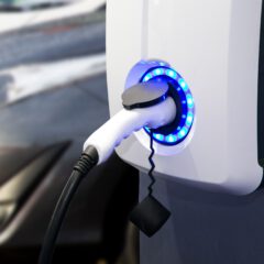 A close-up of an electric vehicle charging station with a white charging plug connected and blue lights glowing around the socket. A blurred car is visible in the background.