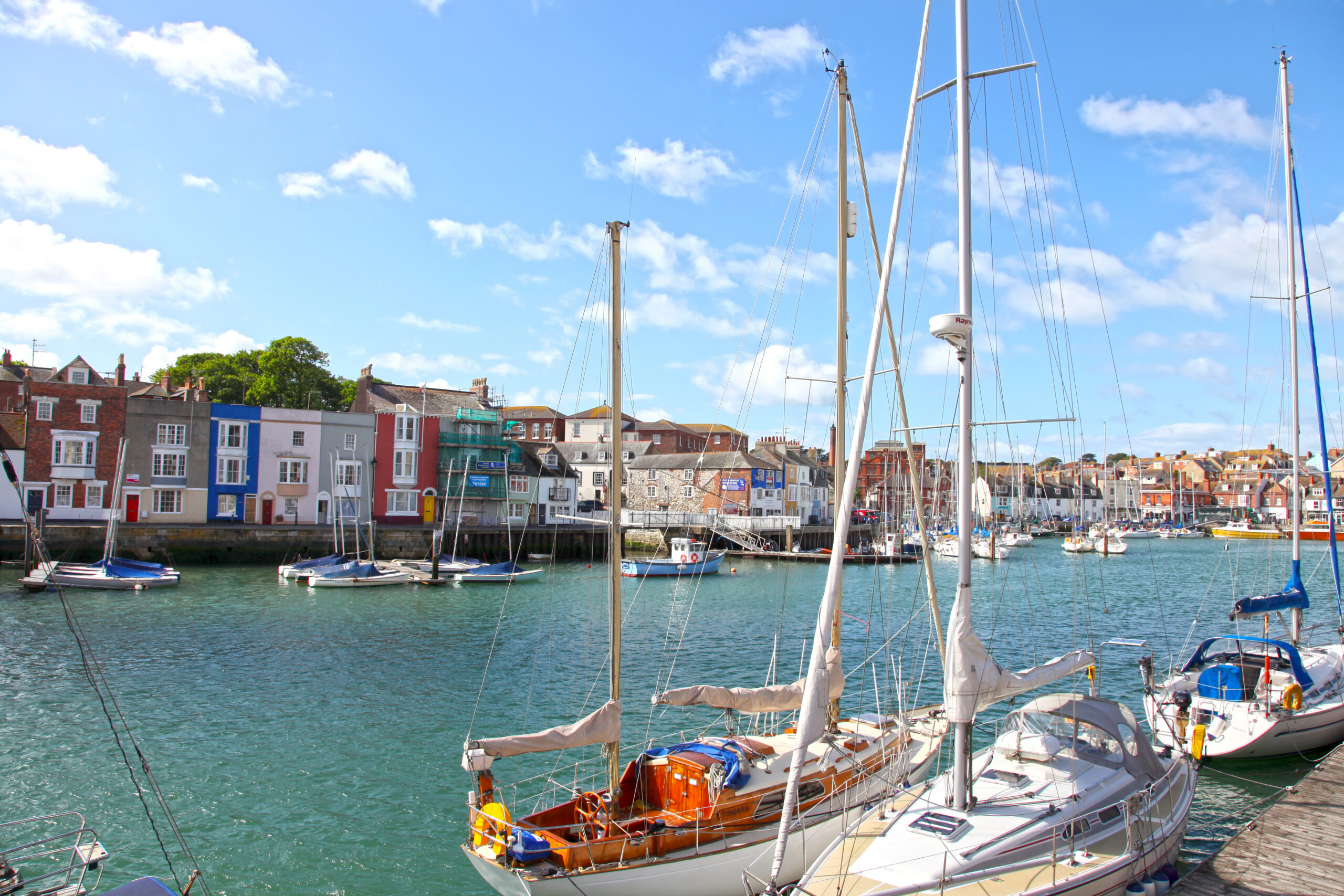 Sailboats docked in a harbor with colorful buildings lining the waterfront under a bright blue sky with scattered clouds. The water reflects the boats and the lively seaside town atmosphere.