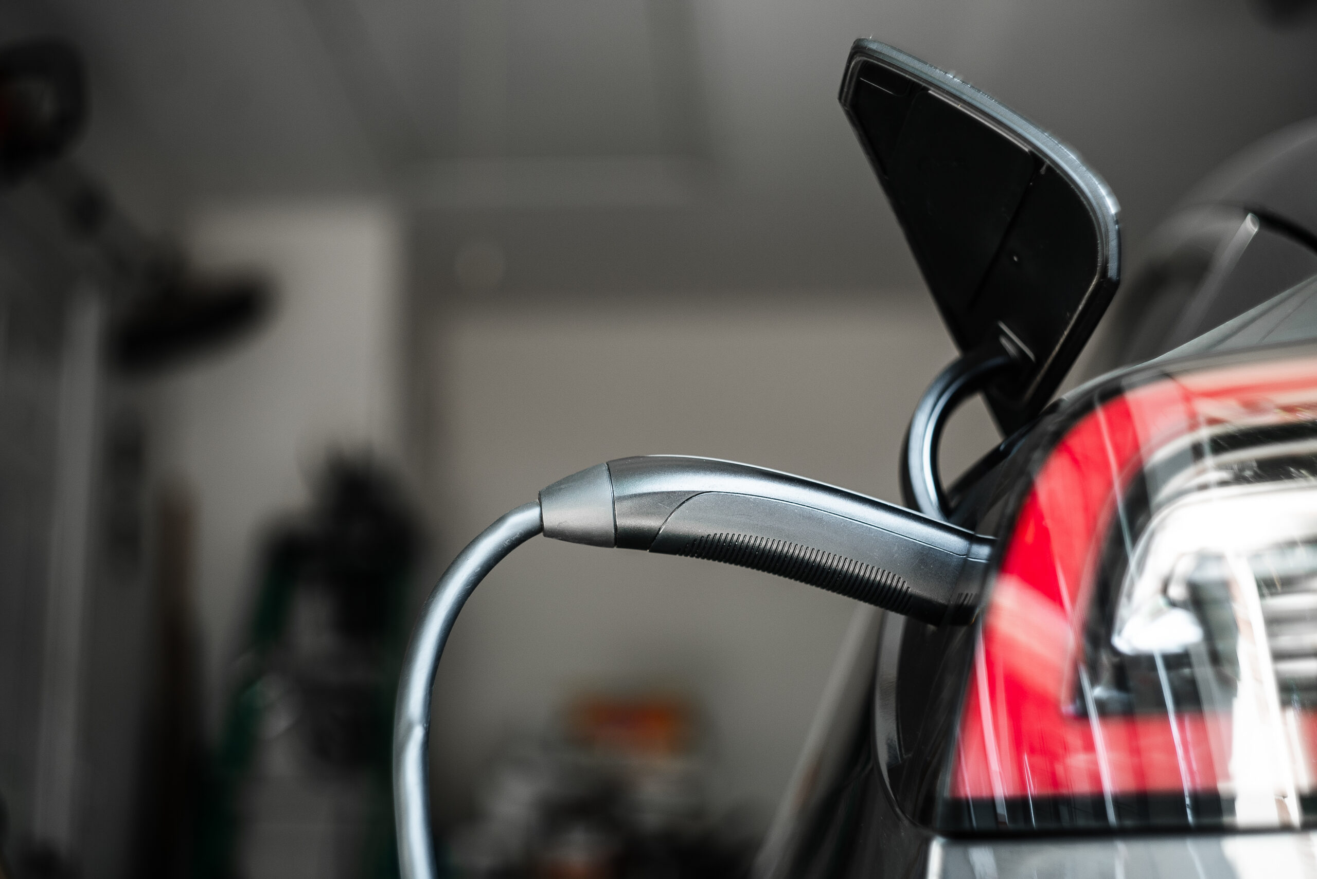 Close-up of an electric car being charged, with the charging cable plugged into the port at the rear of the vehicle. The car's taillight and open charging cover are visible. The background is blurred.