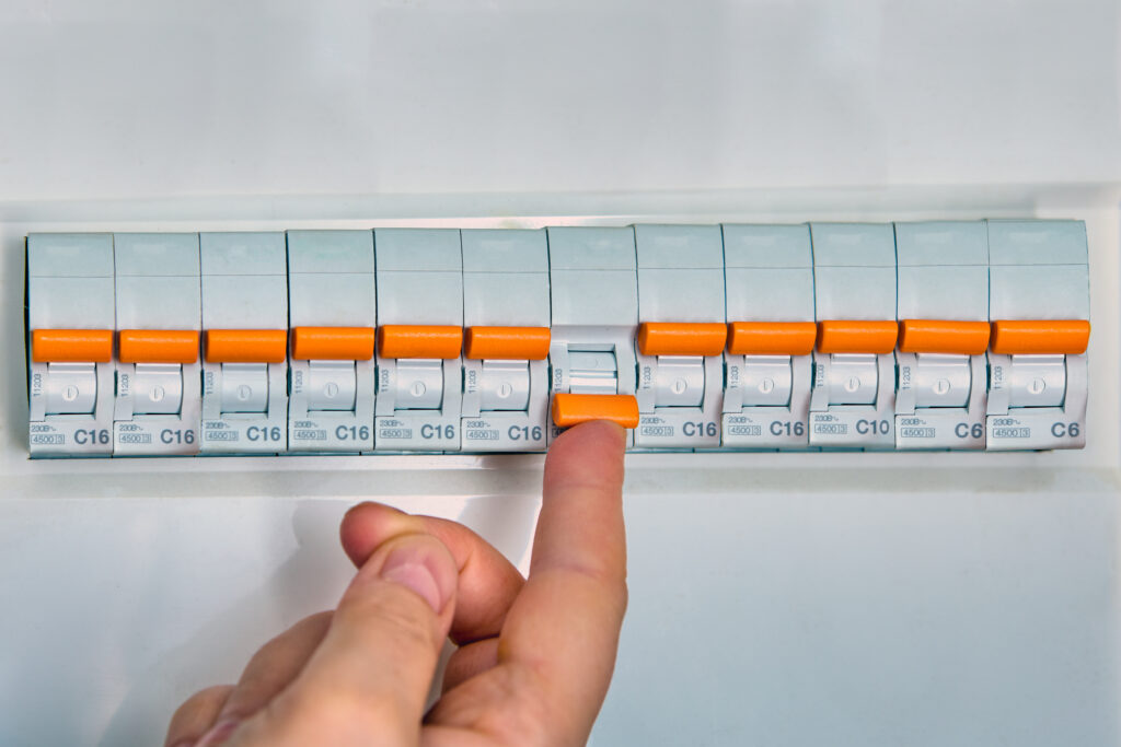 A hand is flipping a switch on an electrical circuit breaker panel with multiple switches, each labeled with numbers and orange handles.