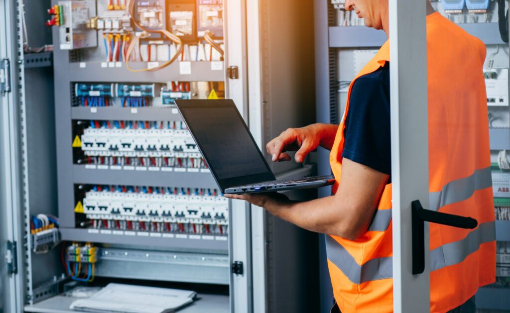 An electrician wearing an orange safety vest uses a laptop while standing in front of an open electrical control panel filled with wiring and circuit breakers.