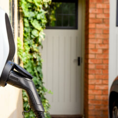 A close-up of an electric vehicle charging point mounted on an exterior wall near a brick house, with a car parked nearby and a white door with windows in the background.