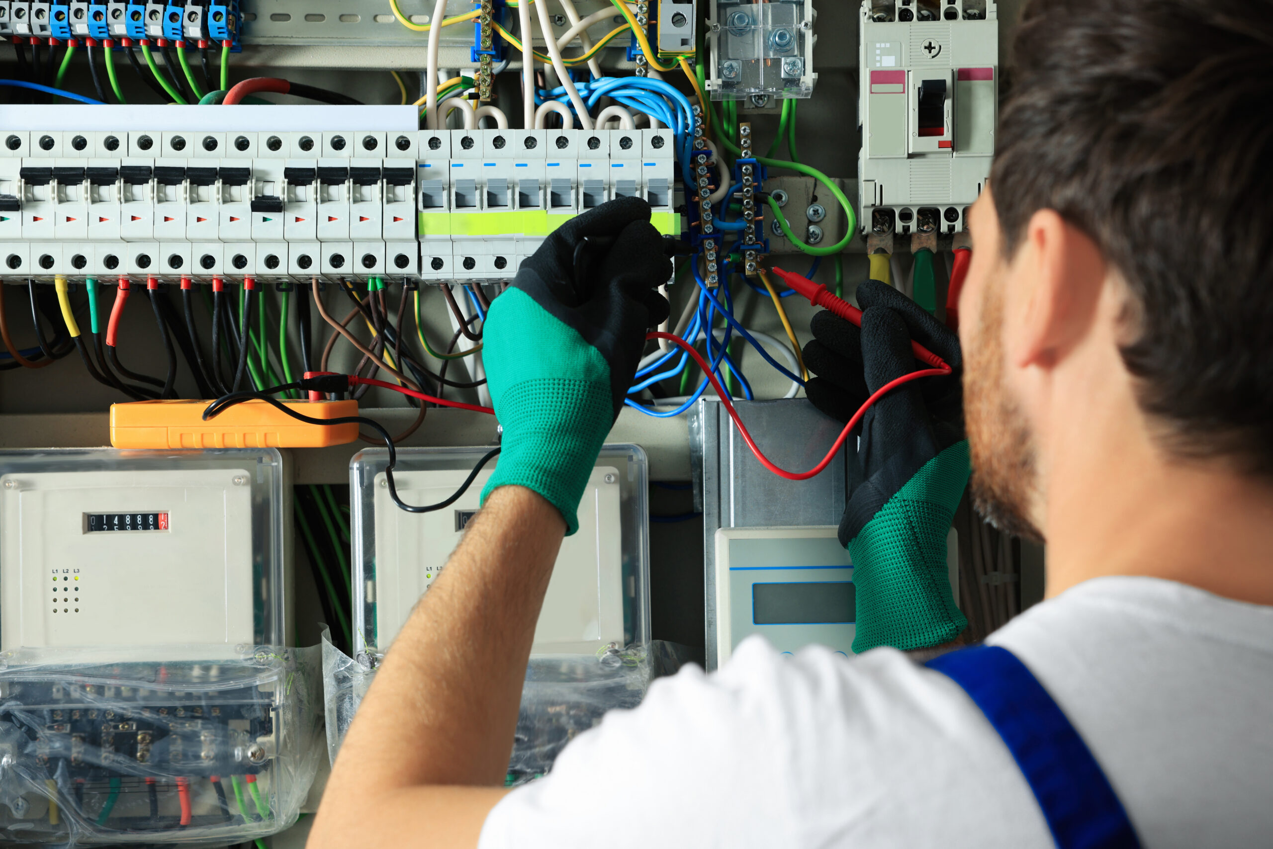 An electrician wearing green gloves uses tools to work on a complex electrical panel filled with colorful wires and circuit breakers.