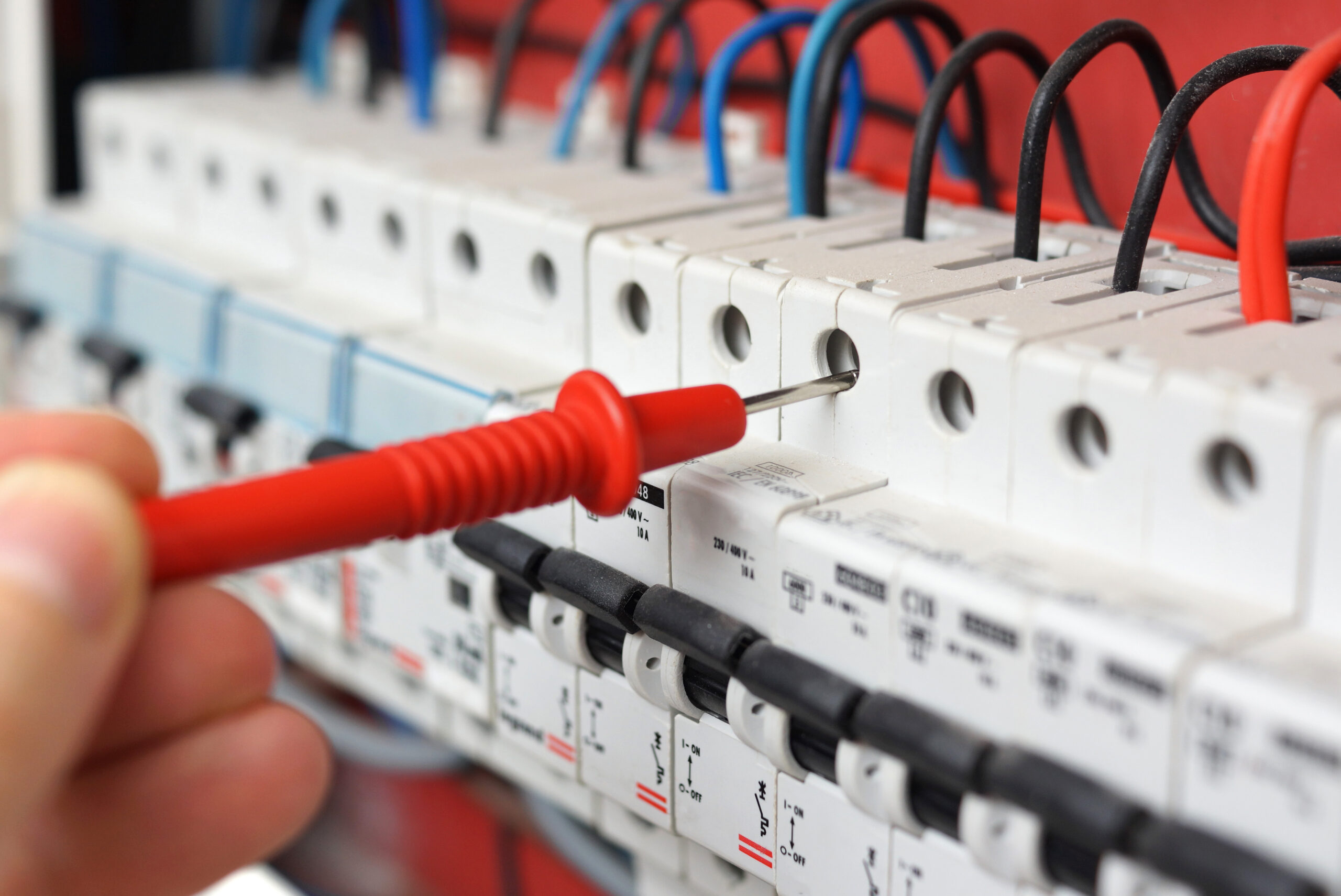 A hand holds a red probe testing a row of electrical circuit breakers with attached wires in an electrical panel.