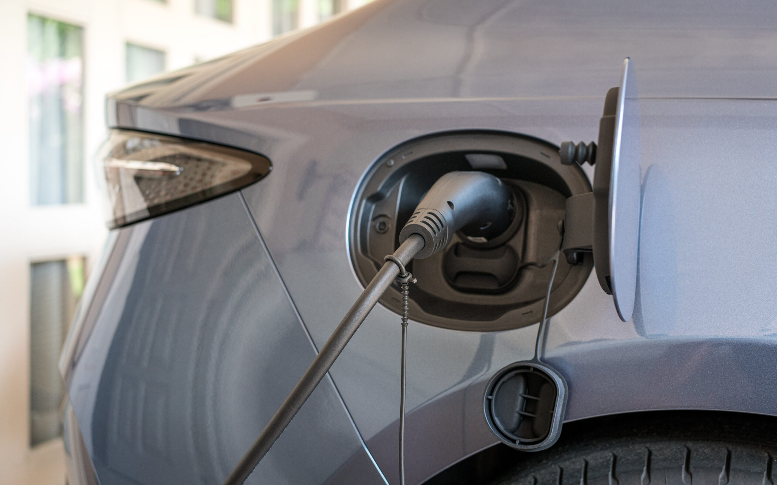 Close-up of an electric car being charged, showing a charging cable plugged into the vehicle’s charging port on the rear side, with the charge port cover open.