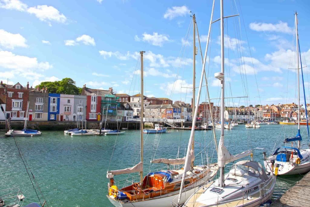 Sailboats are docked in a harbor with colorful waterfront buildings in the background under a bright, blue sky with scattered clouds.