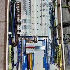 An electrical distribution panel with multiple circuit breakers, organized wiring in various colors, and terminal blocks, mounted on a wall. The wires are neatly arranged along the edges of the panel.