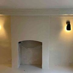 A cream-colored wall with a central, arched fireplace covered by plastic, flanked by two modern black wall sconces emitting soft yellow light; visible is a wooden beam above and a yellow cable on the floor.