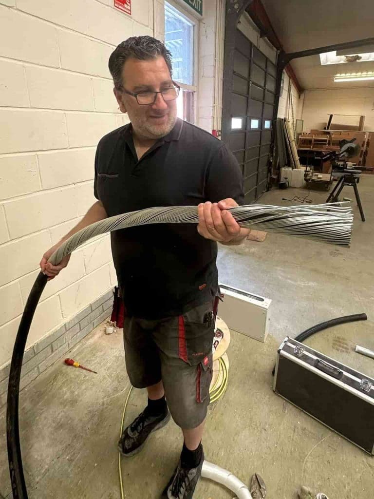 A man wearing glasses and a black polo shirt stands indoors, smiling while holding a thick metal cable. Tools and equipment are scattered on the floor around him in a workshop or industrial setting.
