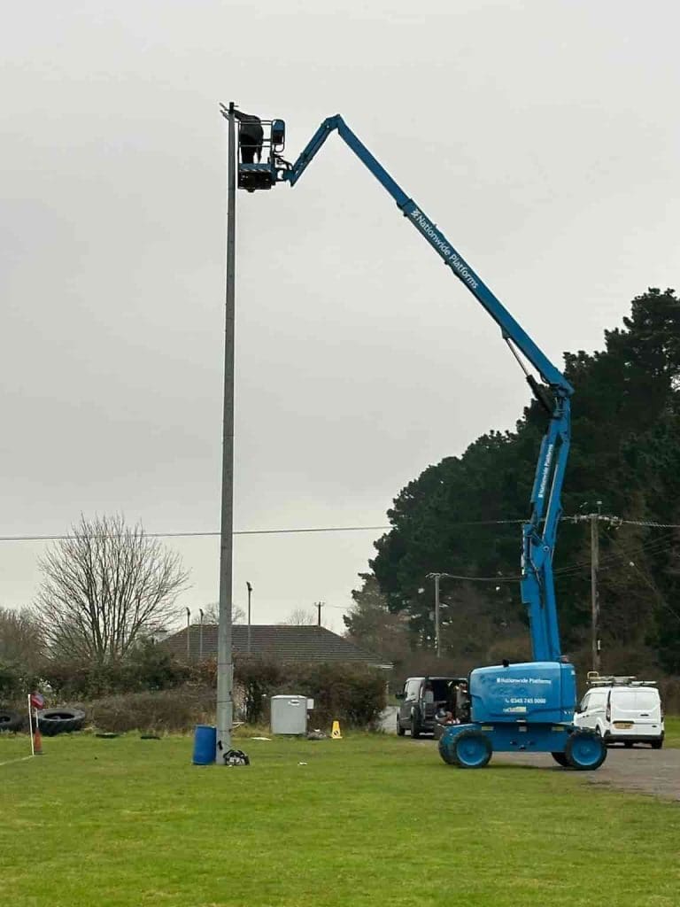 A person in a blue cherry picker lift repairs or inspects a tall light pole on a grassy field, with trees, vehicles, and equipment visible in the background under a cloudy sky.
