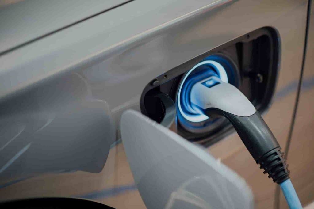 Close-up of an electric car being charged, showing a charging cable plugged into the vehicle’s charging port. The car is silver and the focus is on the connection point.
