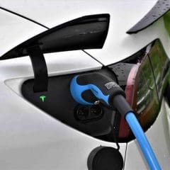 A close-up of a white electric car being charged, with a blue charging cable plugged into the port near the tail light and a green glowing Tesla logo visible.
